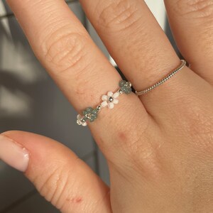 Puede incluir: Un delicado anillo hecho con cuentas de flores blancas y verdes. El anillo se lleva en un dedo.