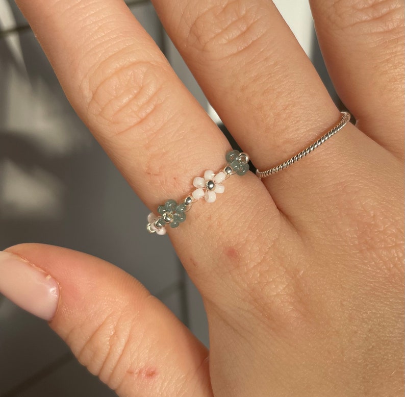 Puede incluir: Un delicado anillo hecho con cuentas de flores blancas y verdes. El anillo se lleva en un dedo.