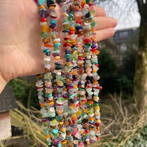 May include: A collection of gemstone bead strands in various colours. The beads are of irregular shapes and include turquoise, orange, purple, green, and red. The gemstones are strung together, creating a vibrant and textured look. The background is blurred.