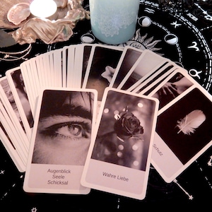 May include: A spread of black and white oracle cards with German text. The cards feature images of an eye, a rose, a feather, and other abstract designs. The cards are laid out on a black cloth with a zodiac symbol pattern.