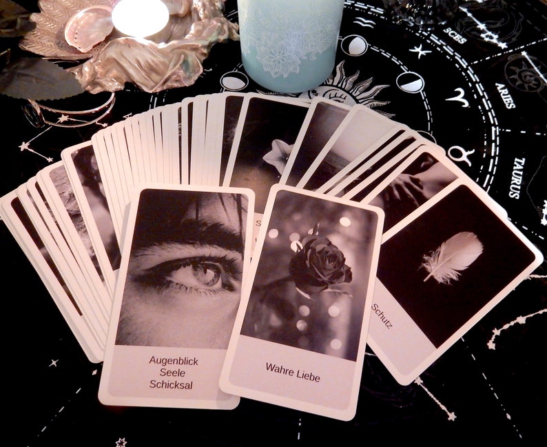 May include: A spread of black and white oracle cards with German text. The cards feature images of an eye, a rose, a feather, and other abstract designs. The cards are laid out on a black cloth with a zodiac symbol pattern.