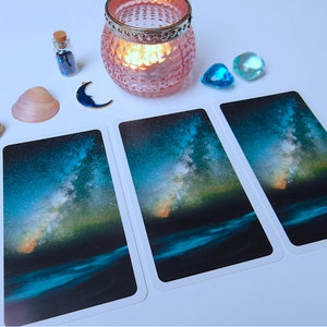 May include: Three tarot cards with a starry night sky and a body of water in the foreground. The sky is a deep blue with a bright, swirling galaxy of stars. The water is a dark blue with a slight ripple. The cards are white with a white border.