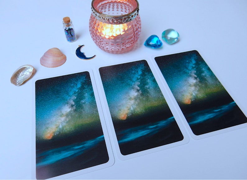 May include: Three tarot cards with a starry night sky and a body of water in the foreground. The sky is a deep blue with a bright, swirling galaxy of stars. The water is a dark blue with a slight ripple. The cards are white with a white border.