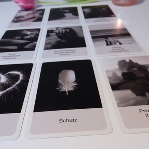 May include: Black and white oracle cards with images and text. The cards include images of a heart, a feather, a castle, and a person in water. The text on the cards includes words like "Taur Samlichk Genus", "Beobachtung Social Media Fotos", "Schutz", "Privatsph&auml; Zu Haus Verste", and "Romantik Ihle Theit".