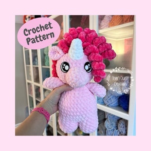Könnte beinhalten: Ein rosa gehäkeltes Einhorn-Amigurumi-Spielzeug mit einem weißen Horn und rosa Wollhaaren. Das Spielzeug hat große schwarze Augen mit weißen Pupillen und eine rosa Nase. Das Spielzeug wird von einer Hand gehalten.