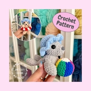 Puede incluir: Un juguete de peluche de foca tejido a ganchillo gris con un sombrero de sol azul claro y una pelota a rayas multicolor. La imagen incluye un recuadro circular que muestra un pequeño oso beige con un salvavidas rojo y blanco y el texto Crochet Pattern.