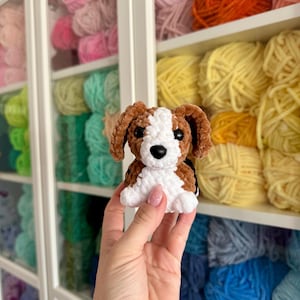 May include: A handmade crochet dog in brown, white, and black. The plush toy has a soft, textured appearance and is sitting. The background shows shelves filled with colorful yarn skeins, suggesting a craft or hobby theme.