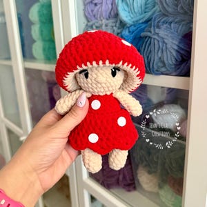 K&ouml;nnte beinhalten: Eine geh&auml;kelte Pilzpuppe mit roter Kappe und einem wei&szlig;en Punktekleid. Die Puppe h&auml;lt eine kleine wei&szlig;e Blume.
