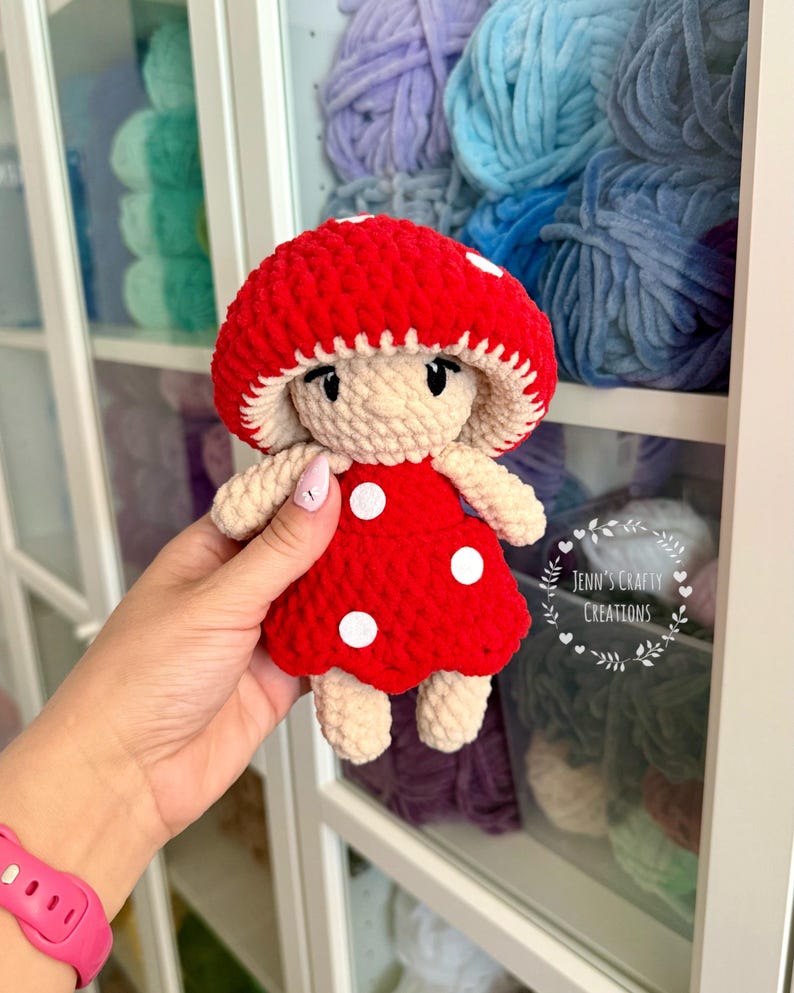 K&ouml;nnte beinhalten: Eine geh&auml;kelte Pilzpuppe mit roter Kappe und einem wei&szlig;en Punktekleid. Die Puppe h&auml;lt eine kleine wei&szlig;e Blume.
