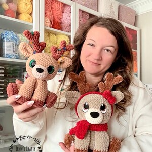 May include: Two handmade crochet reindeer plushies. One is light brown with a red scarf and hat, the other is light brown with a red and green antler decoration. The reindeer have large, expressive eyes and are sitting.