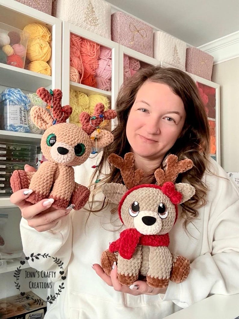 May include: Two handmade crochet reindeer plushies. One is light brown with a red scarf and hat, the other is light brown with a red and green antler decoration. The reindeer have large, expressive eyes and are sitting.