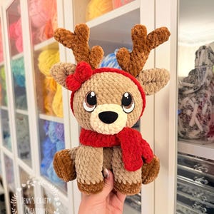 May include: A handmade, plush reindeer toy with brown antlers, a red scarf, and a red bow. The reindeer has large, expressive eyes and is made of soft, textured yarn. The text "Jenn's Crafty Creations" is visible in the lower left corner.