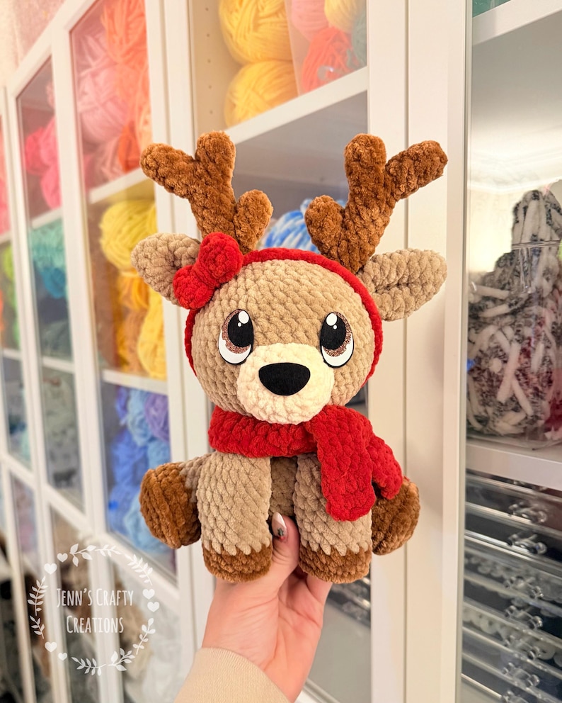 May include: A handmade, plush reindeer toy with brown antlers, a red scarf, and a red bow. The reindeer has large, expressive eyes and is made of soft, textured yarn. The text "Jenn's Crafty Creations" is visible in the lower left corner.