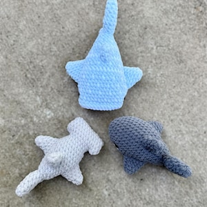 May include: Three crocheted shark toys in shades of blue and gray. The sharks include a light blue whale shark, a gray hammerhead shark, and a dark gray shark. The toys are made of a soft, textured yarn.