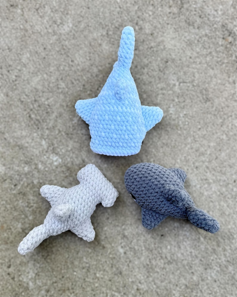 May include: Three crocheted shark toys in shades of blue and gray. The sharks include a light blue whale shark, a gray hammerhead shark, and a dark gray shark. The toys are made of a soft, textured yarn.