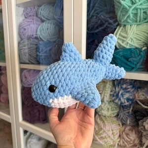 May include: A light blue and white crocheted shark toy with a black eye. The shark is made of soft yarn and has a detailed fin and tail. The background shows shelves filled with colorful yarn.