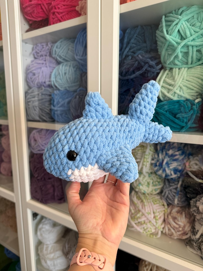 May include: A light blue and white crocheted shark toy with a black eye. The shark is made of soft yarn and has a detailed fin and tail. The background shows shelves filled with colorful yarn.