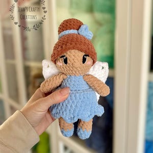 K&ouml;nnte beinhalten: Eine geh&auml;kelte Feenpuppe mit braunen Haaren, einem blauen Kleid und wei&szlig;en Fl&uuml;geln. Die Puppe h&auml;lt eine blaue Schleife in ihren Haaren.