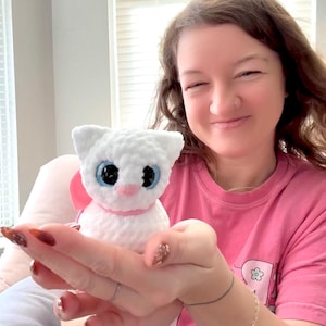 Op de afbeelding: Een handgemaakte witte gehaakte kattenpop met grote blauwe ogen, een roze neus en een roze lint. De kat wordt in de hand van een persoon gehouden. De pop is een zacht, pluche speelgoed.