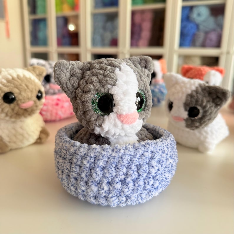 Op de afbeelding: Een handgemaakte gehaakte kattenpop met grijs en wit haar, zittend in een blauwe mand. De kat heeft grote, glinsterende groene ogen en een roze neus. Andere gehaakte dieren staan op de achtergrond.
