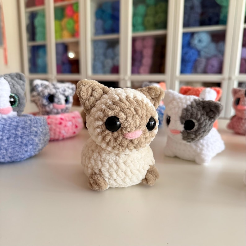 Op de afbeelding: Een verzameling handgemaakte gehaakte kattenbeeldjes in verschillende kleuren. De centrale kat is beige en bruin met grote zwarte ogen en een roze neus. Andere katten zijn wit, grijs en roze. De achtergrond bevat planken gevuld met kleurrijk garen.