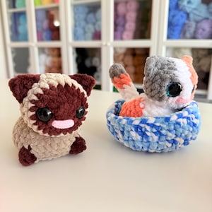 Op de afbeelding: Twee gehaakte dierenfiguren. E&eacute;n is een bruin en tan hondachtig wezen met grote zwarte ogen en een roze mond. De andere is een lapjeskat in een blauw-witte mand. De kat heeft oranje, witte en grijze vacht en grote zwarte ogen.