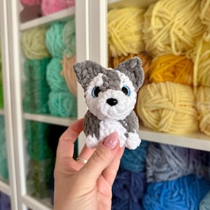 May include: A handmade crochet husky dog with gray, white, and black details, including blue eyes and a black nose. The small plush toy is held in front of a shelf filled with colorful yarn skeins.