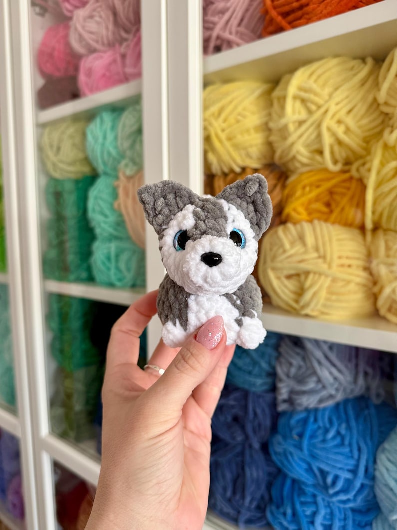 May include: A handmade crochet husky dog with gray, white, and black details, including blue eyes and a black nose. The small plush toy is held in front of a shelf filled with colorful yarn skeins.