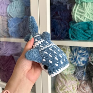 May include: A handmade blue and white crochet whale shark toy with a black eye. The plush toy is held in front of a shelf filled with various shades of blue, green, and purple yarn.