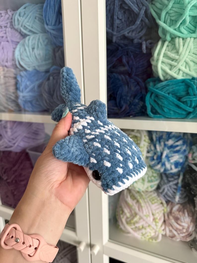 May include: A handmade blue and white crochet whale shark toy with a black eye. The plush toy is held in front of a shelf filled with various shades of blue, green, and purple yarn.