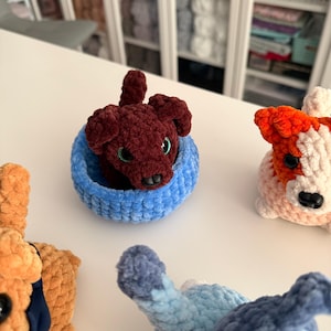 May include: A collection of handmade crochet dog toys in various colors. One dog is a deep burgundy color, nestled in a light blue basket. Other dogs are orange, white, and blue. The toys are soft and plush, with detailed features.