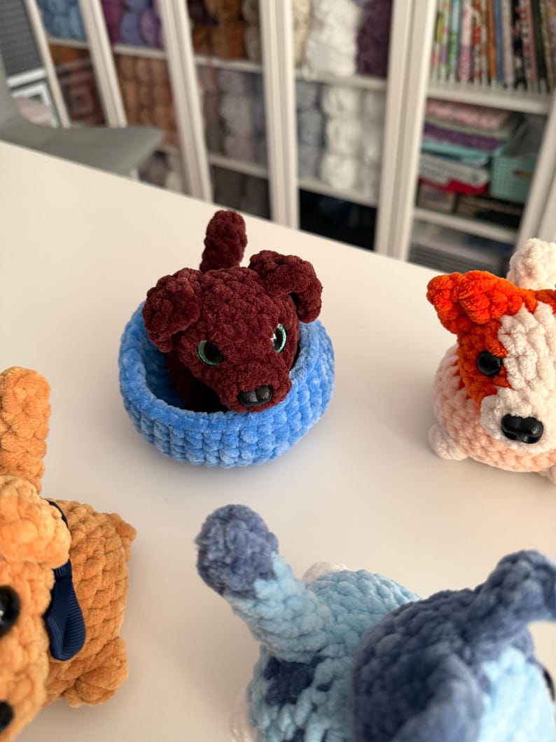 May include: A collection of handmade crochet dog toys in various colors. One dog is a deep burgundy color, nestled in a light blue basket. Other dogs are orange, white, and blue. The toys are soft and plush, with detailed features.