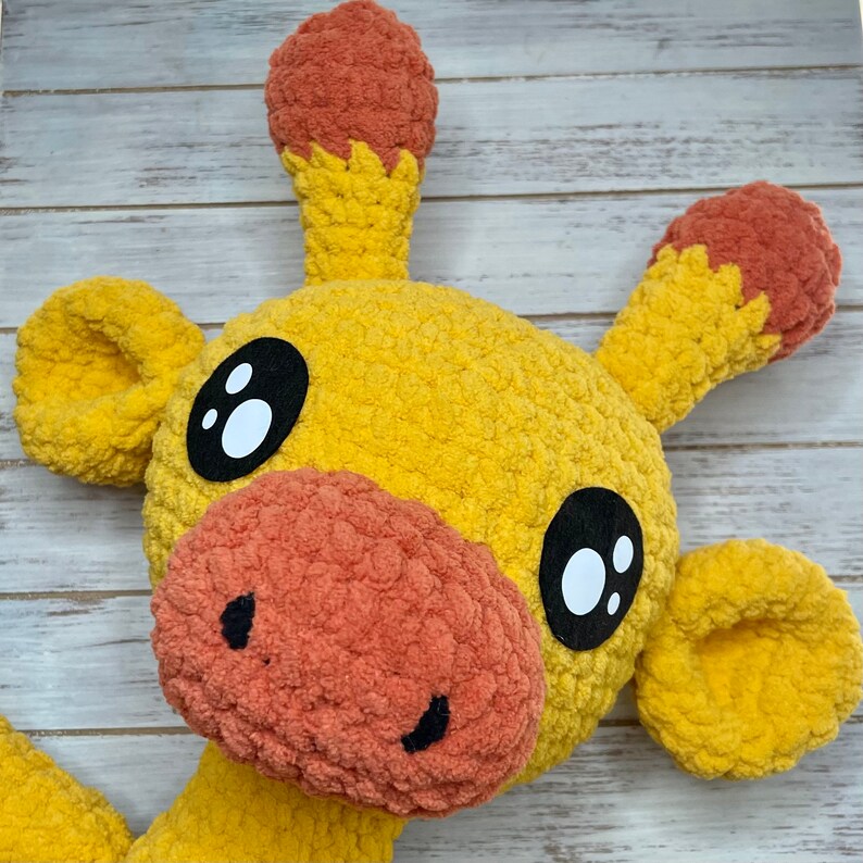 K&ouml;nnte beinhalten: Ein geh&auml;keltes Stofftier-Giraffe mit gelbem K&ouml;rper, orangefarbenen Ohren und schwarzen Augen. Die Giraffe hat eine gro&szlig;e, orange Nase und einen freundlichen Ausdruck.