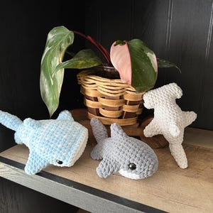 May include: Three crocheted shark plushies in light blue, gray, and white, with black safety eyes, sit on a wooden shelf. A potted plant with green and pink leaves is in the background. The sharks are handmade and have a soft, textured appearance.