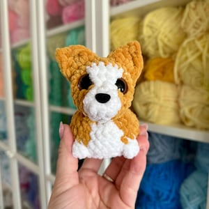 May include: A handmade crochet dog toy in shades of orange, white, and black. The plush toy has large, round eyes and is held in front of a shelf filled with colorful yarn.