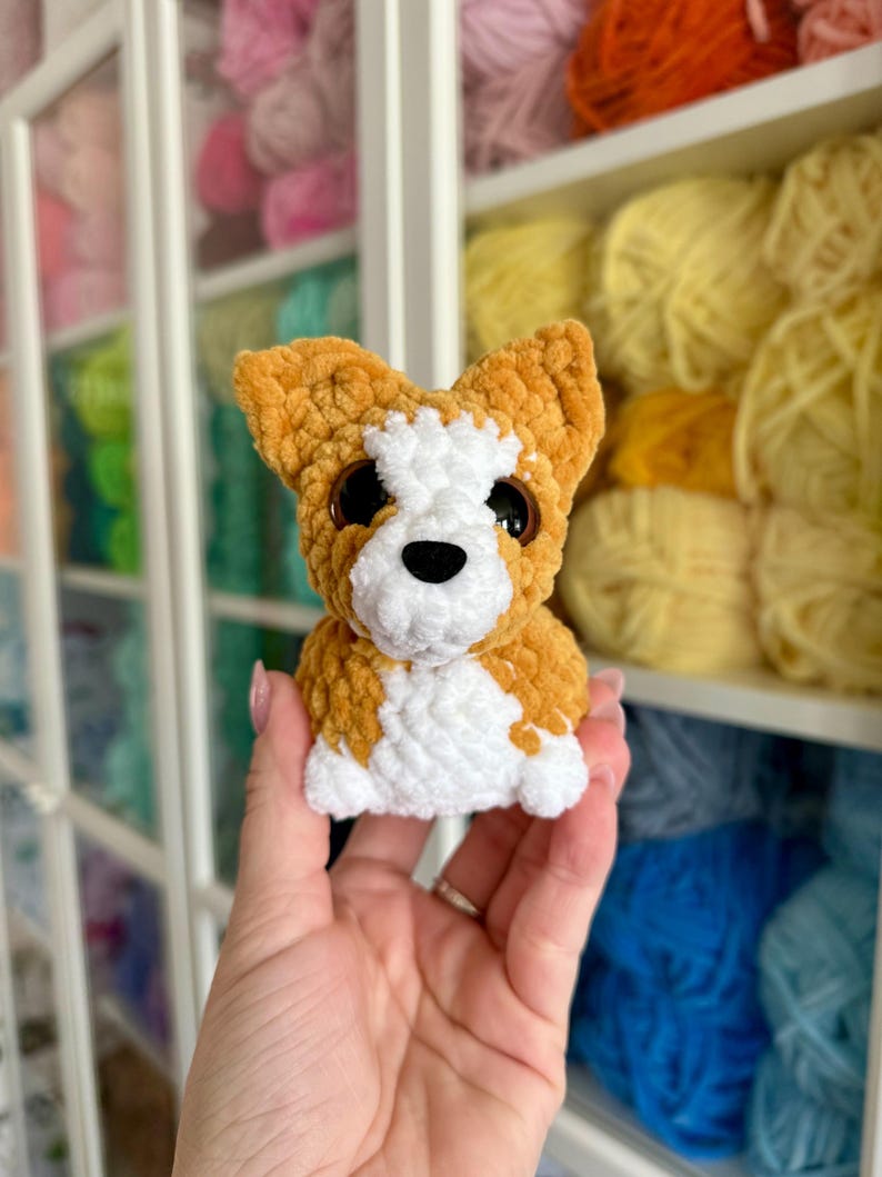 May include: A handmade crochet dog toy in shades of orange, white, and black. The plush toy has large, round eyes and is held in front of a shelf filled with colorful yarn.
