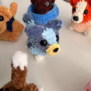 May include: A collection of handmade crochet dog figurines in various colors. The dogs have large black eyes and detailed features. One dog is blue and white, another is orange and white, and a third is brown. The dogs are small and plush.