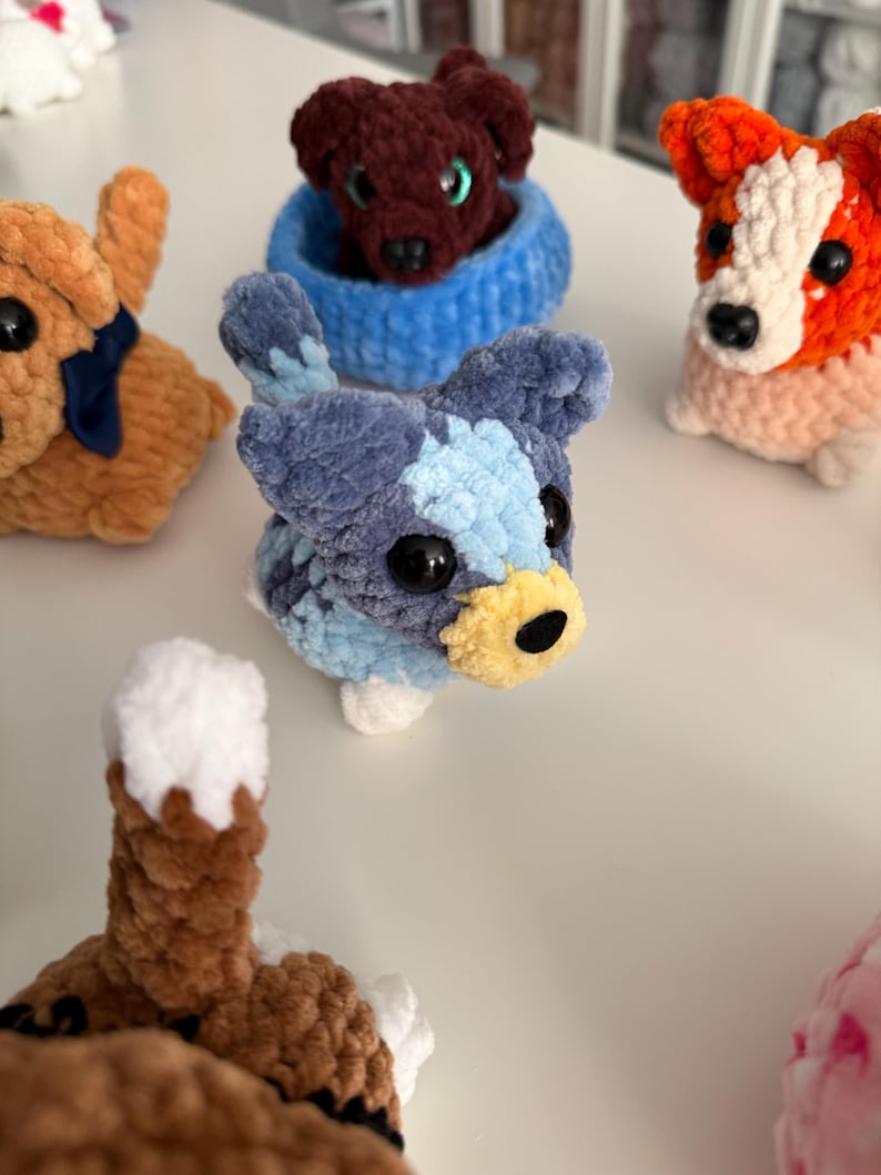 May include: A collection of handmade crochet dog figurines in various colors. The dogs have large black eyes and detailed features. One dog is blue and white, another is orange and white, and a third is brown. The dogs are small and plush.