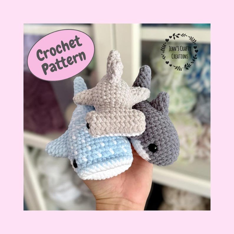 May include: Three crocheted hammerhead shark toys in light blue, gray, and beige. The sharks have black button eyes and white bellies. The image includes a pink oval with the text "Crochet Pattern".