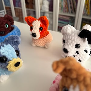 May include: A collection of handmade crochet dog figurines in various colors. The dogs include a brown dog in a blue bed, a red and white dog, a white dog with black spots, a blue and white dog, and a brown dog with a pink bed.