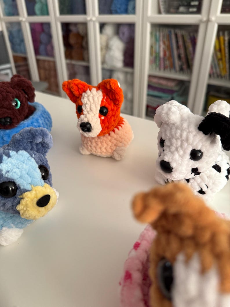 May include: A collection of handmade crochet dog figurines in various colors. The dogs include a brown dog in a blue bed, a red and white dog, a white dog with black spots, a blue and white dog, and a brown dog with a pink bed.