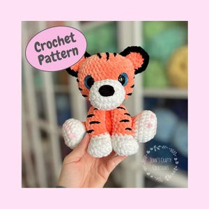 Pode incluir: Um brinquedo de pelúcia de tigre de crochê laranja e preto com patas brancas e olhos azuis. O brinquedo está sendo segurado por uma mão. A imagem inclui o texto "Crochet Pattern".