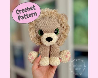 May include: Crocheted lion amigurumi toy with a beige body, brown mane, and green eyes. The toy is sitting in a person's hand.