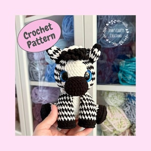 Może przedstawiać: Ręcznie robiona szydełkowa zebra z czarno-białym wzorem na ciele, czarną grzywą i dużymi niebieskimi oczami. Zebra trzyma różowy znak z napisem "Crochet Pattern". Tło zawiera półki z przędzą.