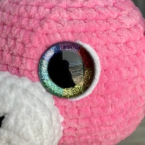 K&ouml;nnte beinhalten: Nahaufnahme eines rosa geh&auml;kelten Stofftiers mit einem gro&szlig;en, glitzernden Regenbogenauge.