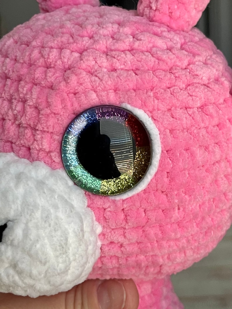 K&ouml;nnte beinhalten: Nahaufnahme eines rosa geh&auml;kelten Stofftiers mit einem gro&szlig;en, glitzernden Regenbogenauge.