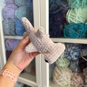 May include: A crocheted hammerhead shark toy in light gray, with a white belly and black eyes. The shark is made of soft, textured yarn. The background shows shelves filled with various colors of yarn.