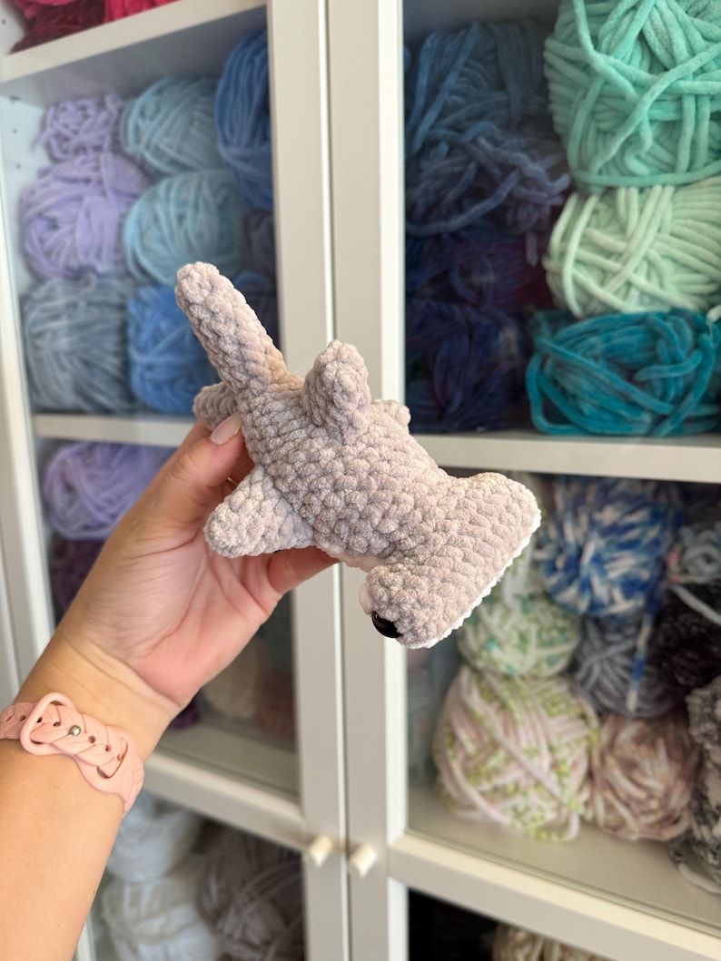 May include: A crocheted hammerhead shark toy in light gray, with a white belly and black eyes. The shark is made of soft, textured yarn. The background shows shelves filled with various colors of yarn.