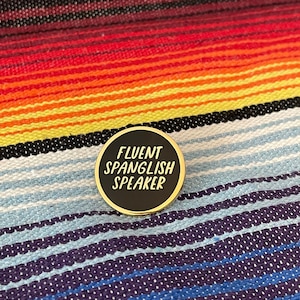 May include: A round, black enamel pin with gold trim. The pin features the text "FLUENT SPANGLISH SPEAKER" in white. The pin is set against a colorful striped textile background.