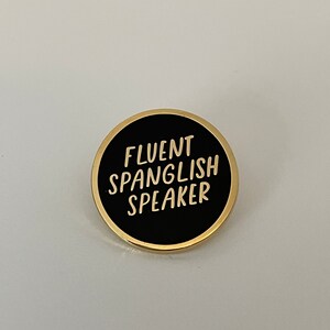May include: A round black pin with gold trim. The pin features the words "FLUENT SPANGLISH SPEAKER" in white, stylized lettering. The pin is set against a white background.